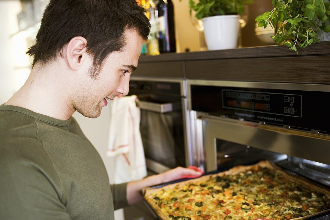 Junger Mann schiebt Blech mit Gemüsepizza in den Ofen