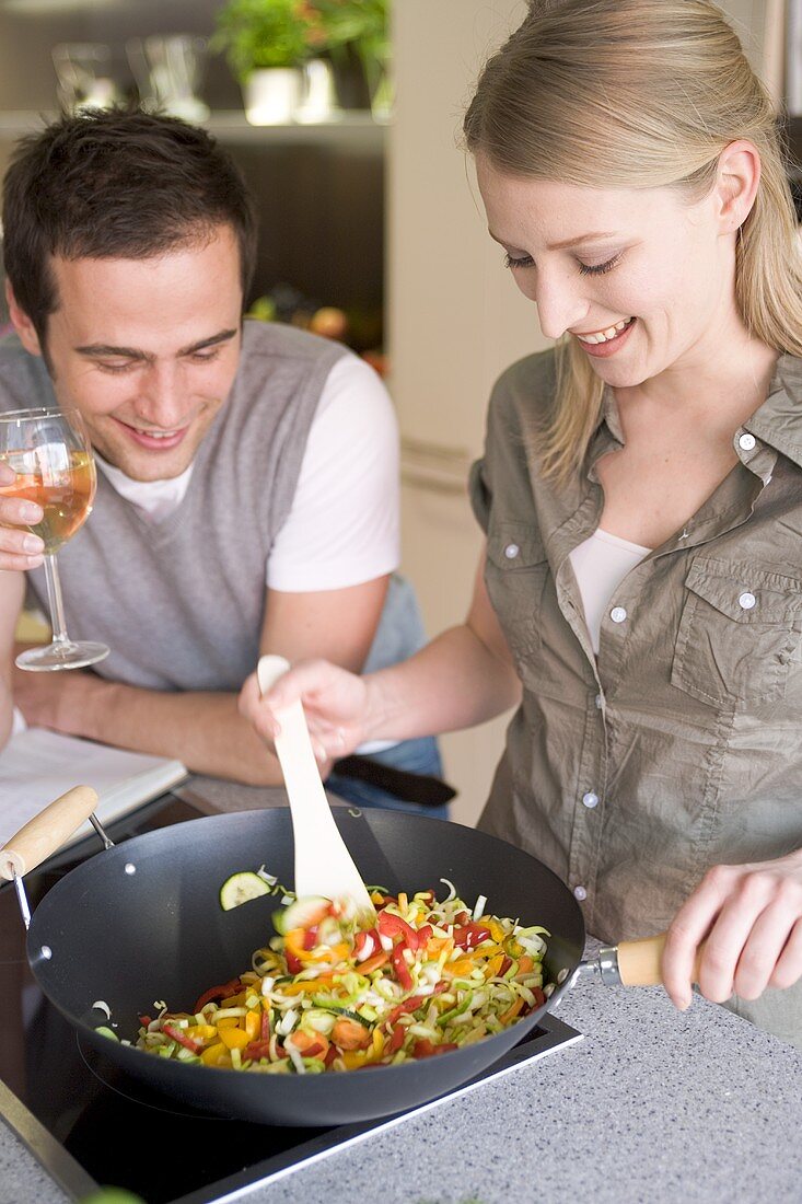 Young woman stirring vegetables in wok, man holding glass of wine