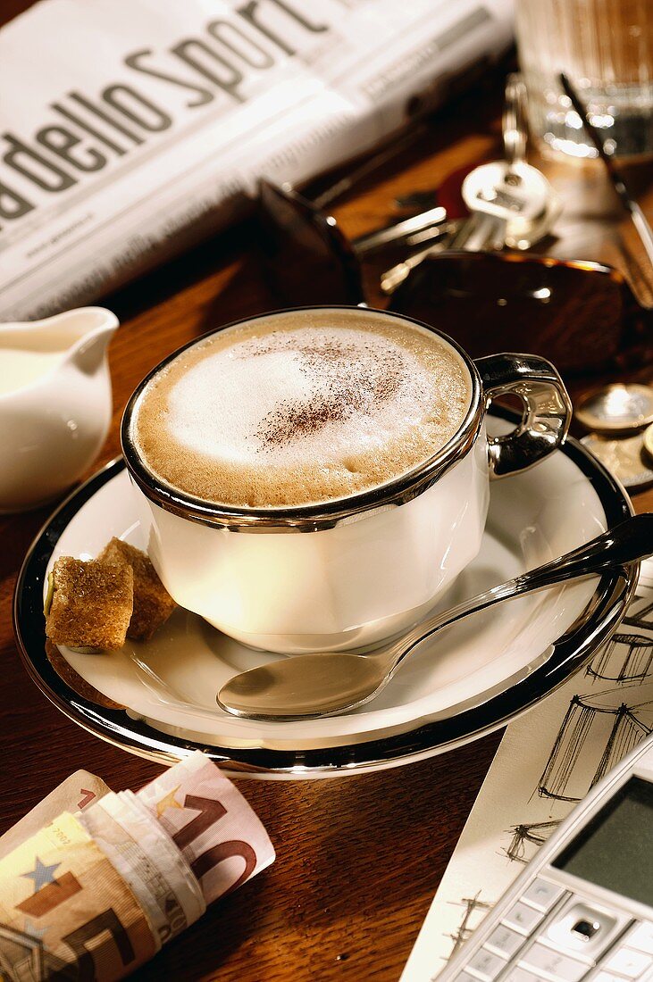 Eine Tasse Cappuccino zwischen Zeitung, Schlüssel und Geld