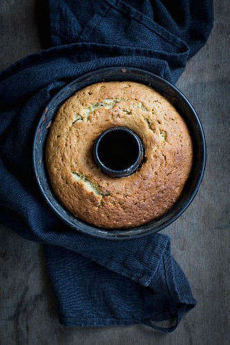 A freshly baked Bundt cake in a baking tin on a dark wooden surface with a grey cloth