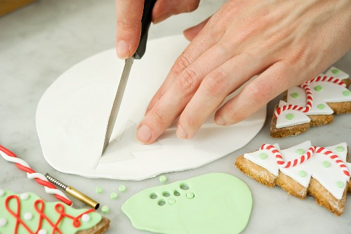 Biscuit decorations being cut out of fondant with decorated Christmas tree biscuits next to it