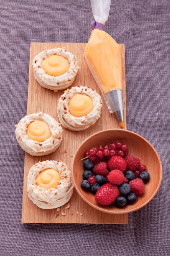 A board with four meringue nests filled with orange cream, next to a piping bag of orange cream and a bowl of fresh berries