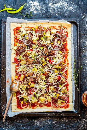 Ungebackene Pizza mit Pulled Pork, Mais, Ananas und Zwiebeln auf Backblech