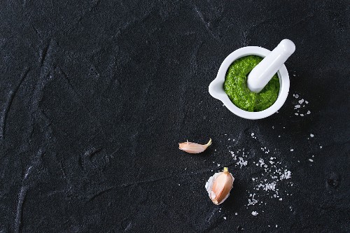 Homemade ramson green pesto sauce in white ceramic mortar with pestle over black textured background