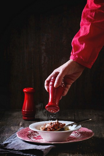 Female hand throws salt from red salt cellar into vintage english plate of homemade pasta pici with Bolognese sauce