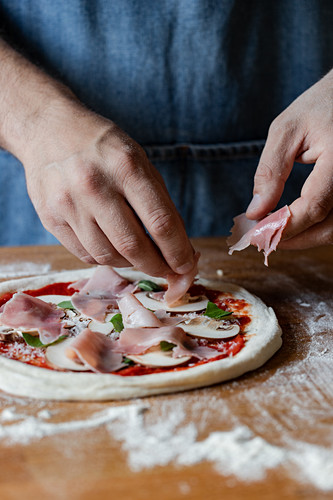 Unrecognizable chef in apron putting tasty ham on raw pizza while cooking on table in kitchen