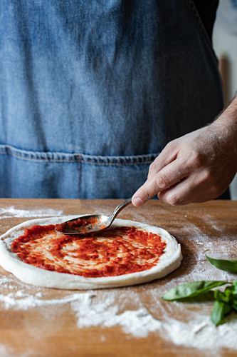 Anonymous chef smearing fresh tomato sauce on raw dough while cooking pizza on table