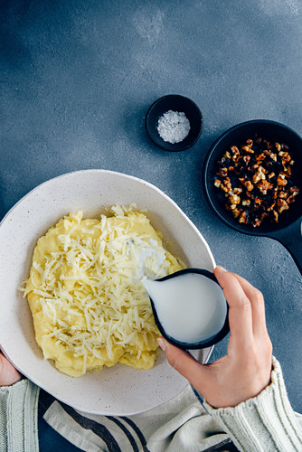 Hands pouring milk from a black ceramic cup on mashed potatoes and grated cheese in a white ceramic bowl