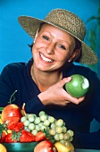Frau mit Hut vor einer Obstschale hält angebissenen Apfel