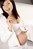 Pregnant woman holding bowl of natural yoghurt & spoon in mouth