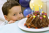 Mischievous boy peeping over table behind birthday cake