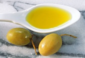 Olive oil on spoon beside two green olives on marble