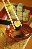 Chopsticks Holding a Single Sushi Roll Over Red Bowl of Sauce