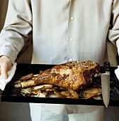 Person holding baking tray with roast leg of lamb