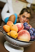 Plums, peaches, apricots in colander, woman in background