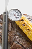 T-bone steak, meat thermometer, corn on the cob, carving fork
