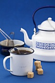 A mug of black tea, sugar cubes, kettle, tea strainer