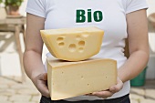 Frau hält zwei grosse Stücke Bio-Käse vor einem Bauernhaus