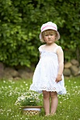 A little girl in a meadow standing next to a straw hat filled with daises