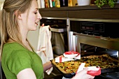 Young woman putting vegetable pizza into oven (on baking tray)