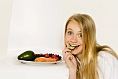 Girl biting into slice of cucumber beside plate of vegetables