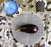 An aubergine on a patterned plate with cutlery, a bowl and a cup on a floral-patterned tablecloth