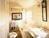 Small bathroom with white wooden walls, fitted cupboard with louver doors, large bathtub and maritime picture on wall