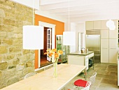 Sonniger Essraum mit Natursteinwand, langer Esstafel mit Metallstühlen, Hängeleuchten und einer zweiten, leuchtendorangen Wand