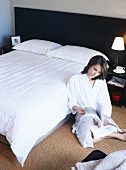 Woman wearing dressing gown sitting reading on floor next to bed in bedroom