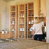 Woman in front of kitchen shelves holding crockery & utensils