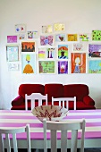 Vor der Wand mit Kinderzeichnungen stehen ein rotes Sofa und ein rosa gestreifter Esstisch