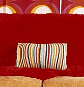 Kissen mit gestreiftem Häkelbezug auf roter Couch vor Tapete im siebziger Jahre Stil