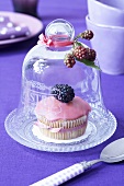 A muffin decorated with pink icing and a blackberry under a cloche