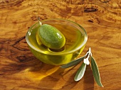 A green olive in a small dish of olive oil, olive leaves