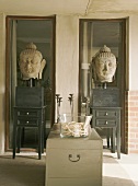 Zwei steinerne Buddha-Köpfe auf Tischchen vor deckenhohen, schmalen Fenstern