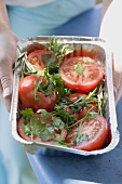Woman holding aluminium dish of tomatoes & herbs for grilling