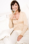 Junge Frau mit einem Glas Weißwein im Bett sitzend