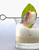 A piece of bass on a skewer above a glass of coconut and vanilla puree with milk foam