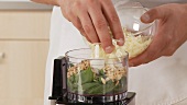 Pine nuts and grated cheese being added to pesto ingredients in a mixer