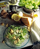Cäsarsalat mit Parmesan auf Glasteller, dahinter Zutaten