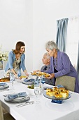 Women Placing Hanukkah Meal on Set Table, Man Sitting at Table