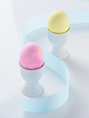Zwei gefärbte Eier in Eierbechern, dazwischen blaue Schleife