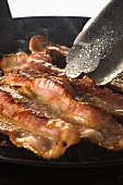 Strips of Bacon Frying in Skillet; Tongs