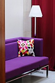 A view of a purple sofa with a colourful cushion and a floor lamp behind