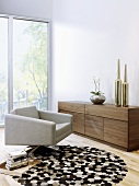 A designer living room - swivel armchair on a round rug and a wooden sideboard in front of a floor-to-ceiling window