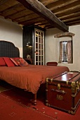 Red leather hope chest in front of a bed with red bed linen under a timber frame ceiling