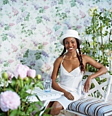 Woman dressed in white on a garden bench in front of a backdrop with flowers and rose trees on it