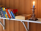 Candlestick with lighted candle and books on a shelf in front of a wood paneled wall