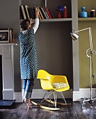 Wohnraum mit gelbem Schaukelstuhl aus Plastik, Stehlampe auf Rollen und einer Frau vor Bücherregal