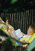 Mädchen liegt mit einem Buch in einer Hängematte im Garten